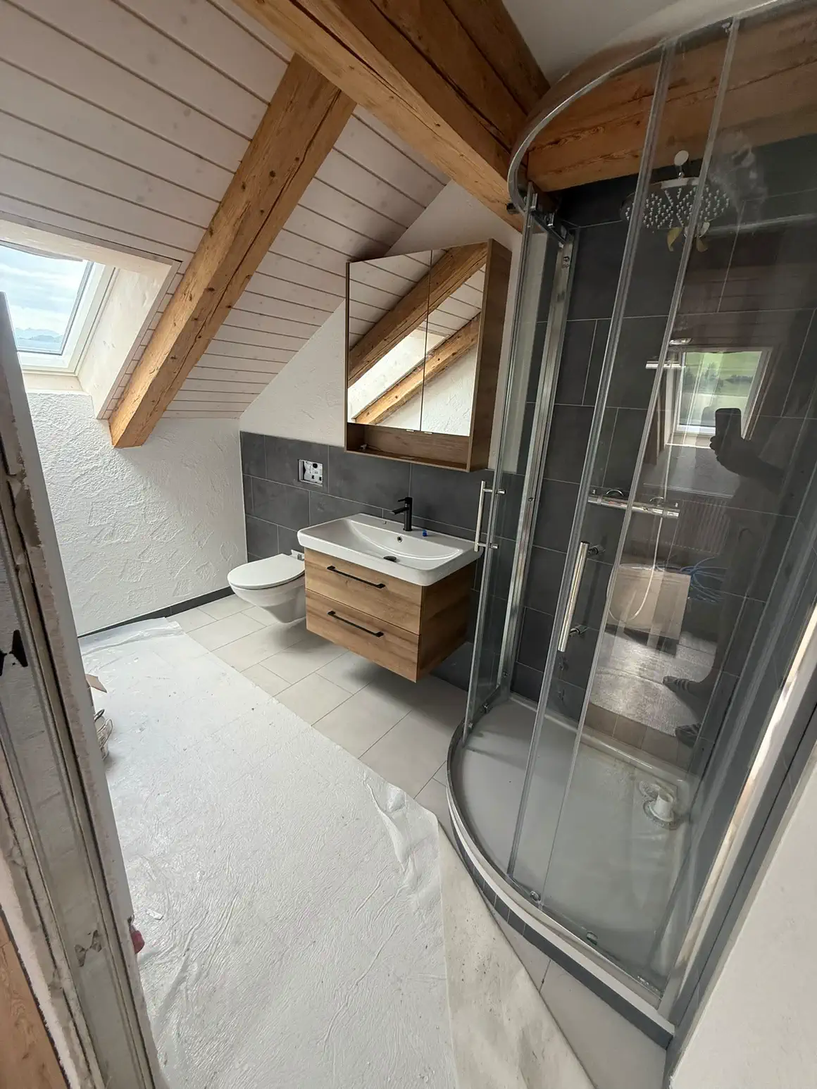 Das fertige Dachgeschoss-Bad mit Viertelkreis-Dusche, Waschtisch und Spiegelschrank unter sichtbarem Altholzbalken
