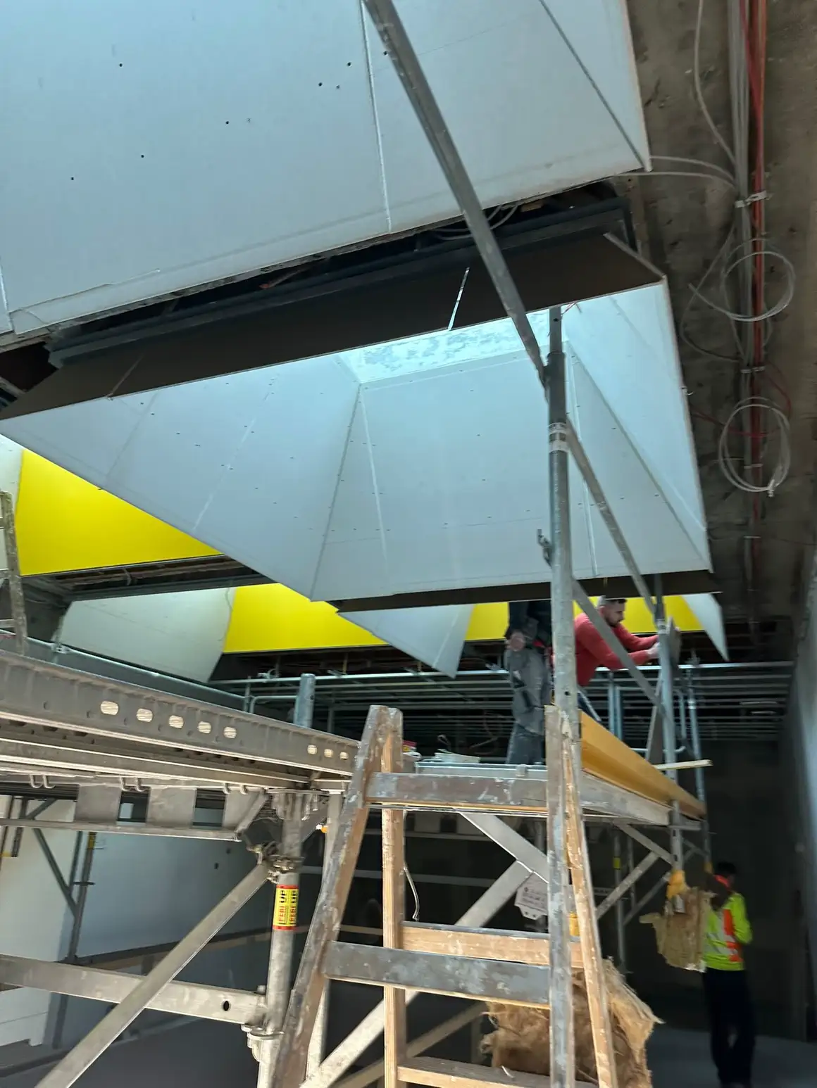 Abgehängte Deckensegel mit angeschrägten Flanken, im Hintergrund die Montage vom Gerüst aus